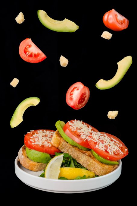 pieces of tomato avocado and cheese levitating above a plate of sourdough toast layered with tomato and avocado and a lemon wedge on the sid