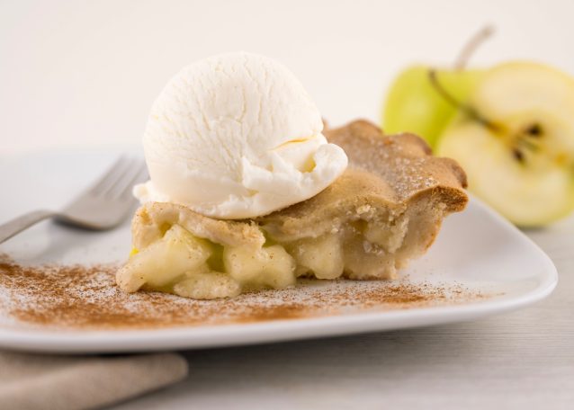 A slice of apple pie with a scoop of vanilla ice cream on top on a plate with sprinkles of cinnamon and green apples in the background