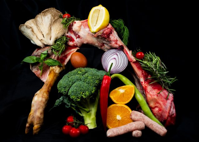 an arrangement of raw vegetables citrus fruit and herbs surrounding a raw beef bone lamb hoof and sausage links