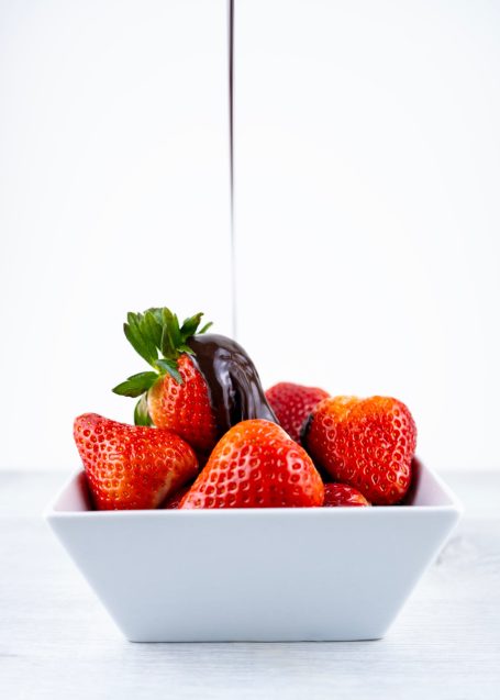 A white square shaped bowl containing brightly coloured strawberries with a chocolate drizzle streaming down from above 