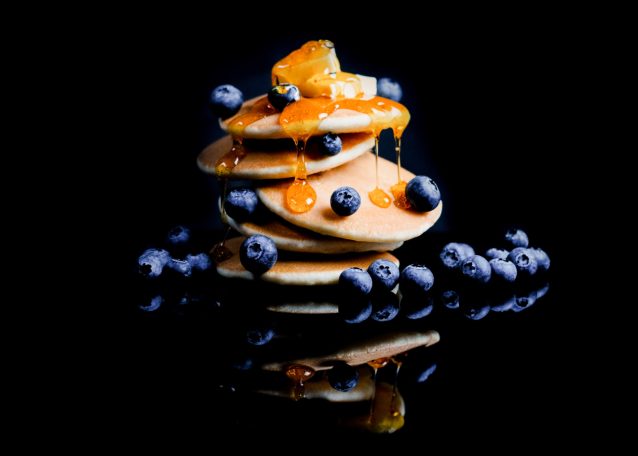A stack of golden pancakes topped with small squares of butter and dripping syrup surrounded by blueberries on a reflective black surface