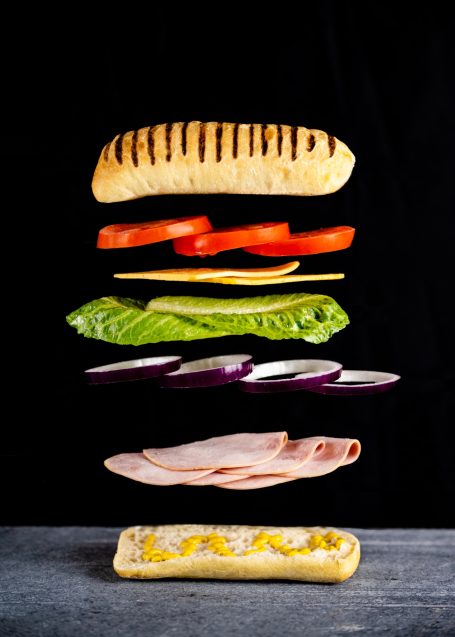 A rustic bun sandwich made with layers of ham onion lettuce cheese and tomato all suspended in mid-air