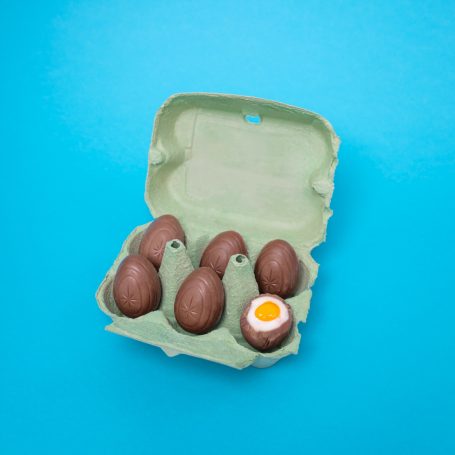 half dozen unwrapped chocolate creme eggs inside of a small egg carton with one chocolate egg broken open to reveal a runny yolk