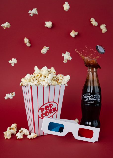 box of popcorn and levitating pieces glass bottle of coke with a popped off cap and splash of cola spraying out and 3-D movie glasses