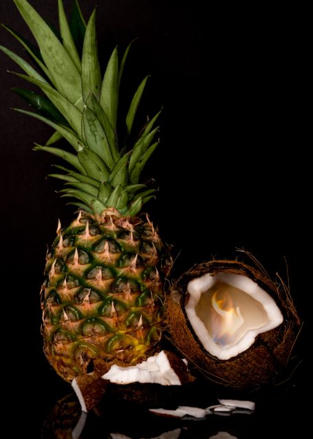 a ripe pineapple beside an open coconut half with a fire inside of the coconut centre