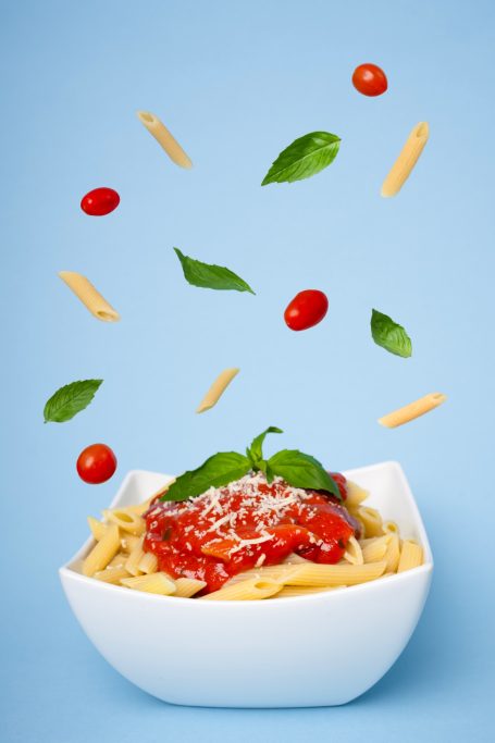 a white bowl of pasta with sauce cheese and basil garnish against a blue background and ingredients levitating above the bowl